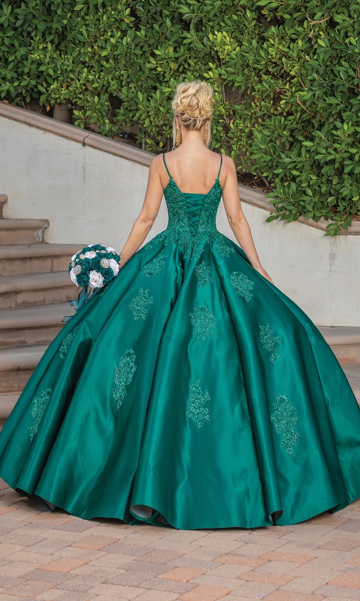 A woman wearing the Quinceanera Dress 1325 by Dancing Queen, a green a-line ball gown with lace applique, stands on a paved area near stone steps and lush greenery, holding a bouquet and facing away from the camera.